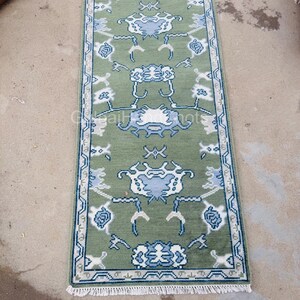 K&ouml;nnte beinhalten: Ein rechteckiger, gr&uuml;ner Teppich mit einem detaillierten Blumenmuster in Wei&szlig; und Blau. Der Teppich hat an beiden Enden einen wei&szlig;en Fransen und einen blauen Rand. Der Text "GulnajHandKnots" ist in der unteren Ecke sichtbar.
