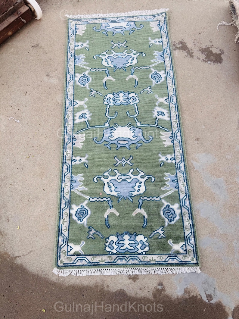 K&ouml;nnte beinhalten: Ein rechteckiger, gr&uuml;ner Teppich mit einem detaillierten Blumenmuster in Wei&szlig; und Blau. Der Teppich hat an beiden Enden einen wei&szlig;en Fransen und einen blauen Rand. Der Text "GulnajHandKnots" ist in der unteren Ecke sichtbar.