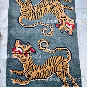 以下が含まれることがあります： 2匹の様式化されたトラが描かれた、長方形のターコイズ色のラグ。トラは金と黒で、口には赤いアクセントがあります。ラグの端はフリンジ加工されています。約150cm x 90cmです。