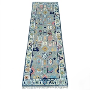 May include: A long, rectangular runner rug featuring a light blue background with a repeating geometric pattern in shades of pink, yellow, navy blue, and white. The rug has a decorative border and short white fringe along both narrow ends.