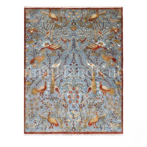 Puede incluir: Una alfombra rectangular con fondo azul claro y un patrón simétrico de aves, árboles y motivos florales estilizados en tonos naranja óxido, crema y marrón, rematada con un borde de color rojo oscuro en los extremos.