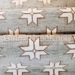 May include: A woven textile runner featuring a repeating pattern of white eight-pointed stars on a light gray background. The stars are outlined in a thin brown thread, and the fabric has a visible grid-like texture throughout the surface.