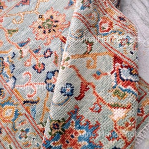 May include: A detailed close-up of a light blue rug with an intricate floral pattern. The rug features a mix of colors, including orange, red, blue, and green, creating a vibrant and textured design. The rug is partially rolled, revealing its underside.