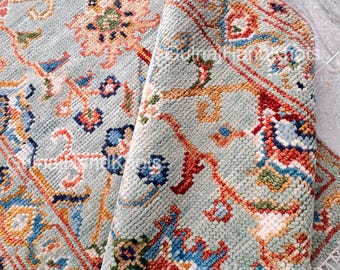 May include: A detailed close-up of a light blue rug with an intricate floral pattern. The rug features a mix of colors, including orange, red, blue, and green, creating a vibrant and textured design. The rug is partially rolled, revealing its underside.