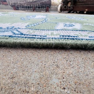 K&ouml;nnte beinhalten: Nahaufnahme eines gr&uuml;n-blau gemusterten Teppichs. Der Teppich hat ein detailliertes Design mit floralen und geometrischen Elementen. Der Rand des Teppichs ist sichtbar und zeigt einen dicken, strukturierten Flor. Der Teppich liegt auf einer Betonoberfl&auml;che.