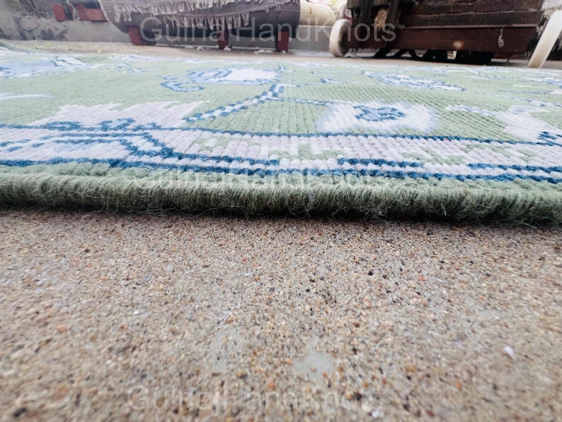 K&ouml;nnte beinhalten: Nahaufnahme eines gr&uuml;n-blau gemusterten Teppichs. Der Teppich hat ein detailliertes Design mit floralen und geometrischen Elementen. Der Rand des Teppichs ist sichtbar und zeigt einen dicken, strukturierten Flor. Der Teppich liegt auf einer Betonoberfl&auml;che.