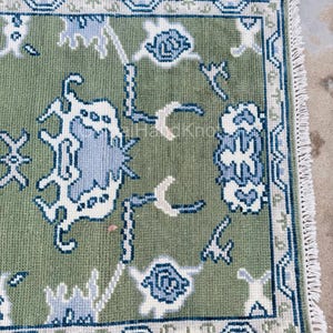 K&ouml;nnte beinhalten: Ein rechteckiger, olivgr&uuml;ner Teppich mit einem detaillierten Blumenmuster in Wei&szlig; und Hellblau. Der Teppich hat einen marineblauen Rand und Fransen auf einer Seite. Der Text "GulnaiHandKnots" ist in den Ecken sichtbar.