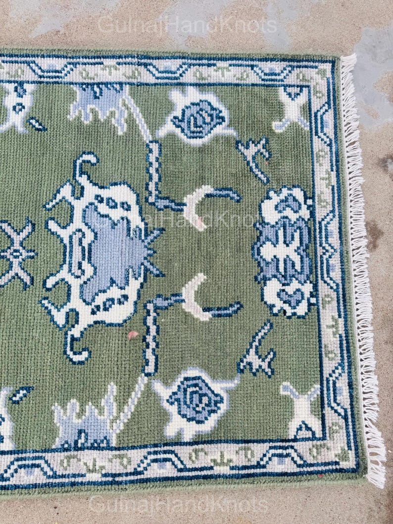 K&ouml;nnte beinhalten: Ein rechteckiger, olivgr&uuml;ner Teppich mit einem detaillierten Blumenmuster in Wei&szlig; und Hellblau. Der Teppich hat einen marineblauen Rand und Fransen auf einer Seite. Der Text "GulnaiHandKnots" ist in den Ecken sichtbar.