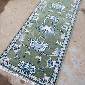 K&ouml;nnte beinhalten: Ein rechteckiger, gr&uuml;ner Teppich mit einem Blumenmuster in Wei&szlig; und Blau. Der Teppich hat einen blauen Rand und einen wei&szlig;en Fransen an den kurzen Enden. Der Text "GulnajHandKnots" ist sichtbar.