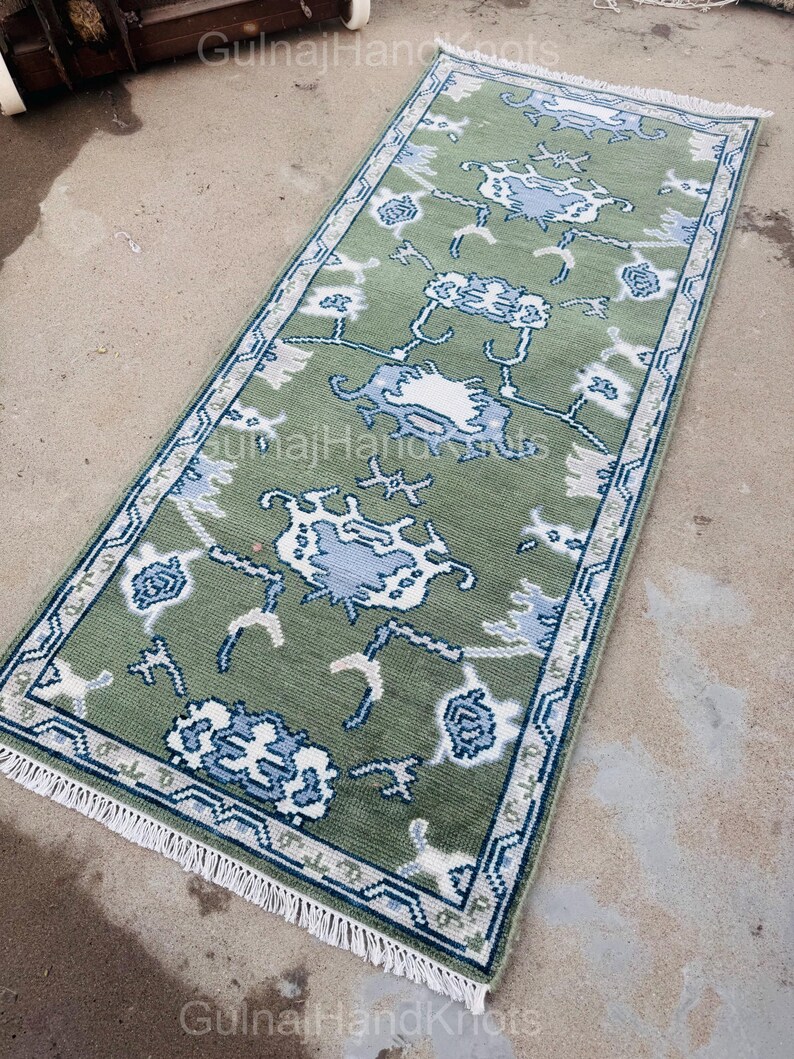 K&ouml;nnte beinhalten: Ein rechteckiger, gr&uuml;ner Teppich mit einem Blumenmuster in Wei&szlig; und Blau. Der Teppich hat einen blauen Rand und einen wei&szlig;en Fransen an den kurzen Enden. Der Text "GulnajHandKnots" ist sichtbar.