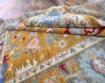 May include: A rolled-up, intricately patterned rug with a warm color palette. The rug features a mustard yellow field with floral and geometric designs in shades of blue, red, and cream. The border is a mix of gray, blue, and cream.