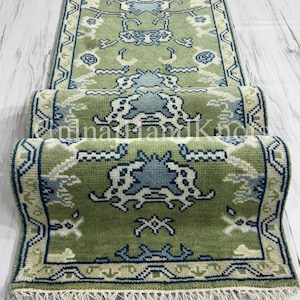 May include: A long, rectangular runner rug featuring a sage green background with intricate, symmetrical patterns in light blue, cream, and dark blue. The rug has a decorative border and a fringed edge at the bottom, with the text "Oina HandKnots" visible.