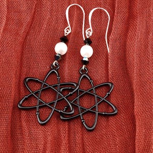 May include: A pair of black atom-shaped earrings with white pearl beads and black accents. The earrings hang from silver-colored ear wires, set against a textured red background. The earrings are a unique accessory.
