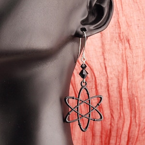 May include: A black atomic symbol earring with a silver hook and two black faceted beads. The earring is displayed on a black mannequin head against a peach-colored fabric background.
