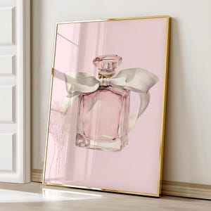 Op de afbeelding: Een ingelijste kunstdruk met een aquarel illustratie van een roze parfumfles met een wit lint. De fles heeft een goudkleurige dop en staat tegen een zachtroze achtergrond. De lijst is goudkleurig.