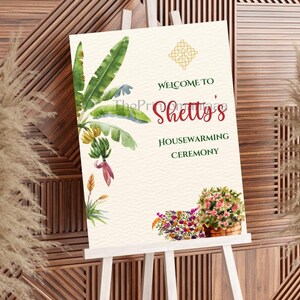 May include: A housewarming ceremony sign on a white easel. The sign features watercolor illustrations of banana leaves, flowers, and text that reads "Welcome to Shetty's Housewarming Ceremony."