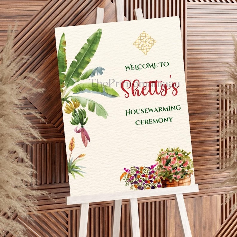 May include: A housewarming ceremony sign on a white easel. The sign features watercolor illustrations of banana leaves, flowers, and text that reads "Welcome to Shetty's Housewarming Ceremony."