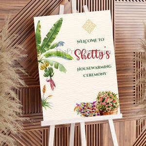May include: A housewarming ceremony sign on a white easel. The sign features watercolor illustrations of banana leaves, flowers, and text that reads "Welcome to Shetty's Housewarming Ceremony."