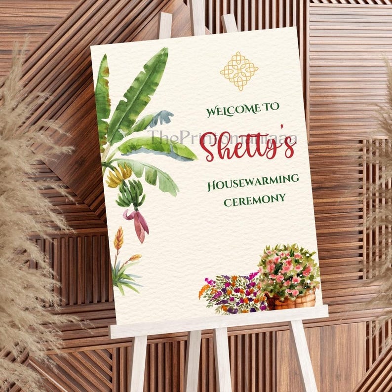 May include: A housewarming ceremony sign on a white easel. The sign features watercolor illustrations of banana leaves, flowers, and text that reads "Welcome to Shetty's Housewarming Ceremony."