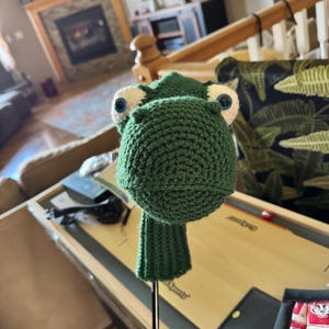 Pode incluir: Uma capa para a cabeça do taco de golfe verde, feita de crochê, em forma de sapo. O sapo tem olhos grandes, redondos e azuis com detalhes brancos e uma cabeça arredondada e texturizada. A capa é projetada para caber em um taco de golfe.