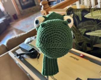May include: A green crocheted golf club head cover in the shape of a frog. The frog has large, round, blue eyes with white accents and a textured, rounded head. The cover is designed to fit over a golf club.