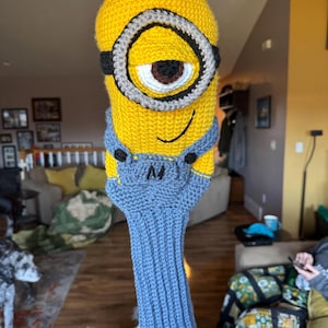 May include: A yellow and blue knitted golf club head cover shaped like a Minion character. The cover features a large eye, black and gray goggles, and blue overalls. The cover is designed to protect golf clubs.