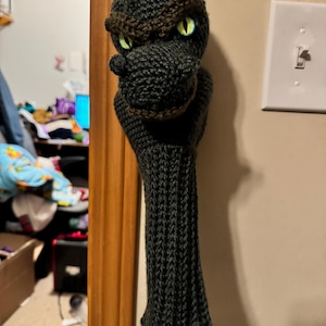 May include: A handmade, dark gray knitted golf club head cover shaped like a dragon's head. The dragon has brown accents, green eyes, and a long, textured body. The cover is designed to protect golf clubs.