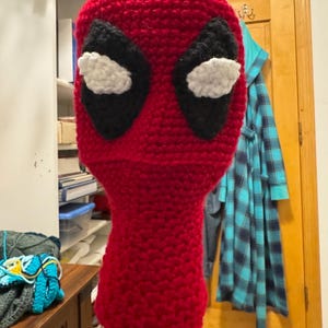 Könnte beinhalten: Eine rote gehäkelte Maske mit schwarzen Augenformen und weißen Augenakzenten. Die Maske ist so konzipiert, dass sie Kopf und Hals bedeckt. Die Maske wird auf einem Ständer ausgestellt. Die Maske ist handgefertigt.