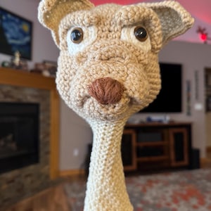 May include: A beige, crocheted dog head cover for a golf club. The dog has large, dark eyes, a brown nose, and floppy ears. The cover is designed to fit over the club head, providing protection and a playful aesthetic. The dog's head is detailed with textured yarn.