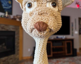 May include: A beige, crocheted dog head cover for a golf club. The dog has large, dark eyes, a brown nose, and floppy ears. The cover is designed to fit over the club head, providing protection and a playful aesthetic. The dog's head is detailed with textured yarn.