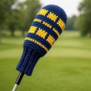Könnte beinhalten: Eine marineblaue und gelb gehäkelte Golfschlägerhaube. Die Haube hat eine gerippte Manschette und ein Muster aus gelben Quadraten und Linien auf marineblauem Hintergrund. Der Golfschläger befindet sich vor einem verschwommenen grünen Hintergrund.