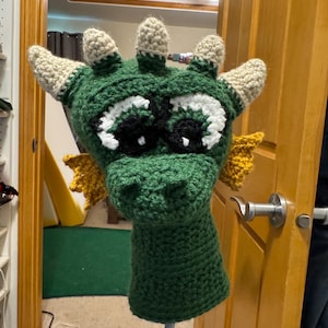 Op de afbeelding: Een groene gehaakte drakenhoofdbeschermer met witte en zwarte ogen, beige hoorns en gele accenten. De drakenhoofdbeschermer is ontworpen om over een golfclub te passen. Het handgemaakte item wordt op een blauwe paal getoond.