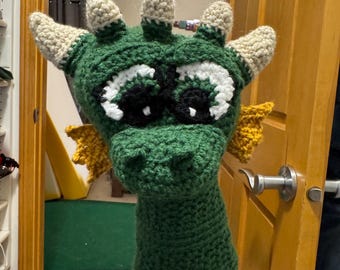 May include: A green crocheted dragon head cover with white and black eyes, tan horns, and yellow accents. The dragon head cover is designed to fit over a golf club. The handmade item is displayed on a blue pole.