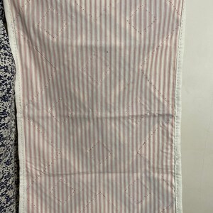 May include: A rectangular quilted fabric with a white border. The interior features a pattern of vertical red and white stripes, with diamond-shaped stitching. The fabric is held up by a person.