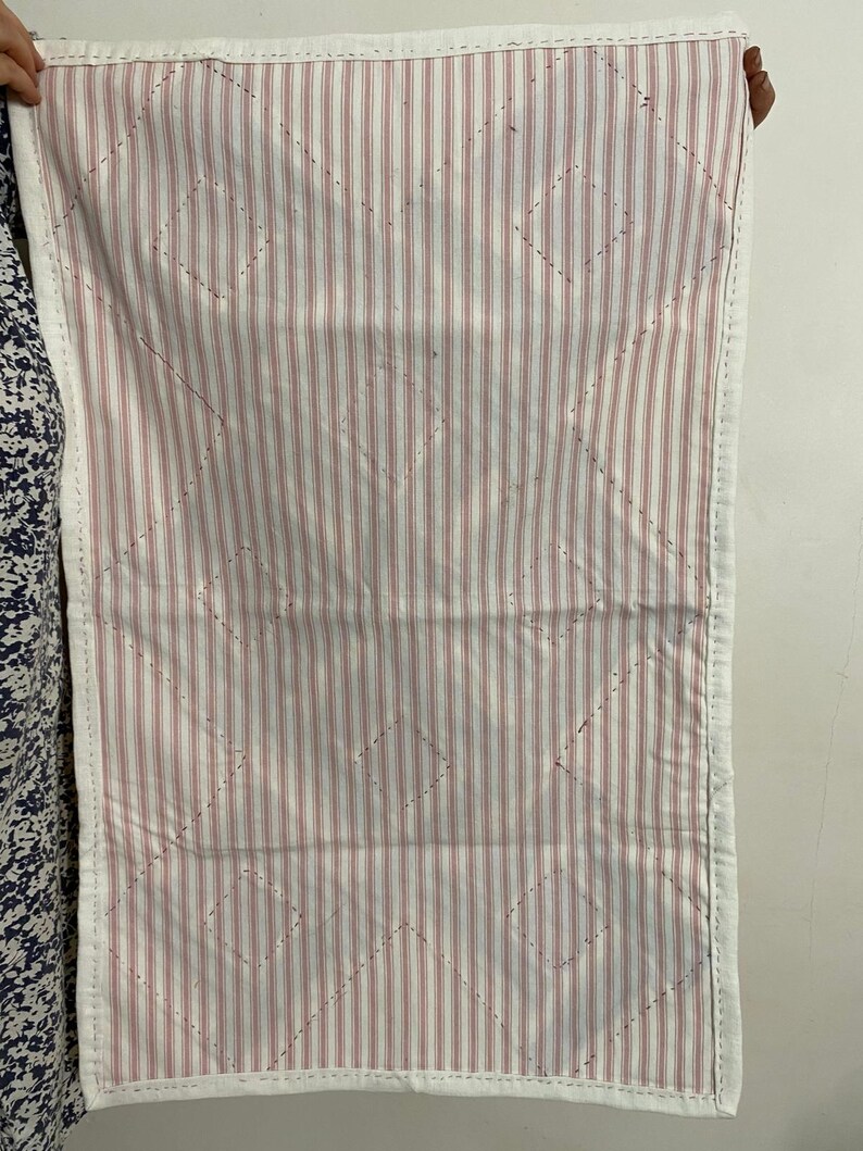 May include: A rectangular quilted fabric with a white border. The interior features a pattern of vertical red and white stripes, with diamond-shaped stitching. The fabric is held up by a person.
