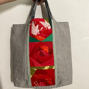 May include: A grey and white striped tote bag with a patchwork rose design. The central panel features a vibrant red rose composed of fabric pieces, bordered by green and light green trim. The bag has grey handles.