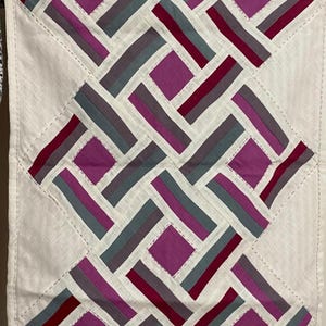May include: Handmade textile art piece with a geometric design. The artwork features a white background with a repeating pattern of rectangles and squares in shades of purple, grey, and burgundy. The edges are finished with a stitched border.