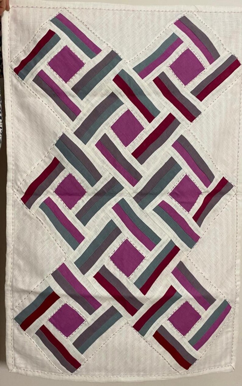May include: Handmade textile art piece with a geometric design. The artwork features a white background with a repeating pattern of rectangles and squares in shades of purple, grey, and burgundy. The edges are finished with a stitched border.