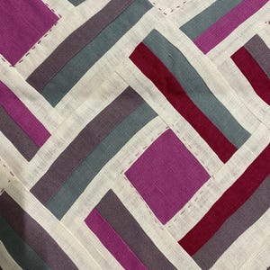 May include: Close-up of a textile with a geometric pattern. The design features alternating rectangles and squares in shades of purple, grey, and burgundy, set against a cream-coloured background. The pieces are stitched together with a contrasting thread, creating a patchwork effect.