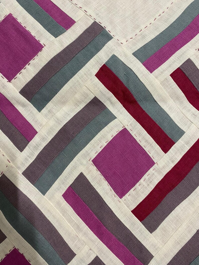 May include: Close-up of a textile with a geometric pattern. The design features alternating rectangles and squares in shades of purple, grey, and burgundy, set against a cream-coloured background. The pieces are stitched together with a contrasting thread, creating a patchwork effect.