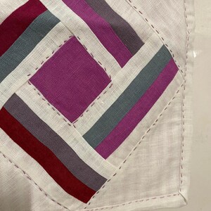 May include: Close-up of a handmade textile with a geometric design. The design features a central square and diagonal stripes in shades of burgundy, grey, purple, and white. The edges are finished with a stitched border.