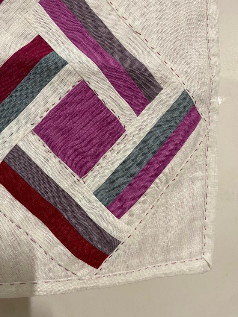 May include: Close-up of a handmade textile with a geometric design. The design features a central square and diagonal stripes in shades of burgundy, grey, purple, and white. The edges are finished with a stitched border.