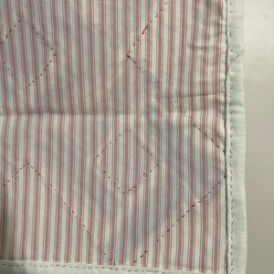 May include: Close-up of a quilted fabric with a white border. The fabric features vertical red and white stripes with a diamond pattern stitched across the surface. The border is a solid white fabric.