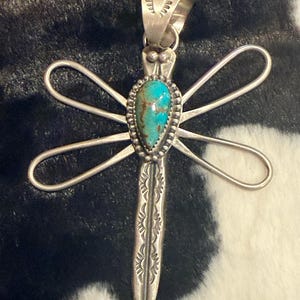 May include: A silver dragonfly pendant featuring a teardrop-shaped turquoise stone with brown matrix in the center. The wings are crafted from smooth silver wire, and the body is decorated with stamped sunburst patterns and a beaded silver border.