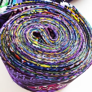 May include: Rolled fabric with a vibrant mix of colors, including purple, yellow, and blue. The fabric appears to be patterned with various designs. The roll is tightly wound, showcasing the layers of material.