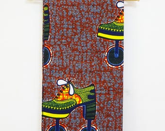 May include: A rectangular piece of fabric featuring a repeating pattern of stylized shoes on wheels. The background is a deep red with small white and blue square motifs. The shoes are green, yellow, and orange with white laces and black wheels.