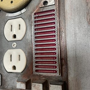 May include: Close-up of a metallic panel with two white electrical outlets. A rectangular section features a series of red, horizontal bars. The panel has a weathered, industrial aesthetic with various geometric shapes and textures.