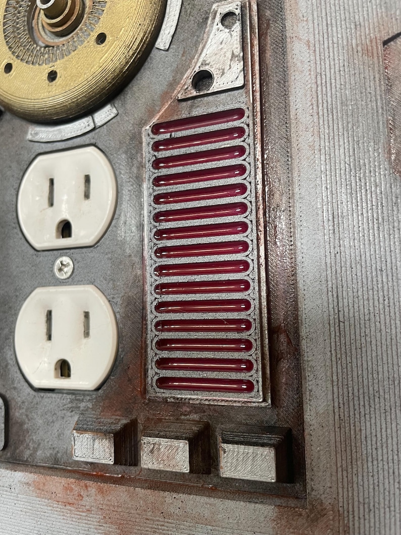 May include: Close-up of a metallic panel with two white electrical outlets. A rectangular section features a series of red, horizontal bars. The panel has a weathered, industrial aesthetic with various geometric shapes and textures.