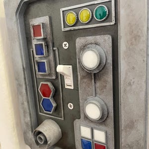 May include: A gray panel with various buttons, switches, and lights. The panel features a toggle switch labeled "ON", multiple colored lights, and several white and colored buttons. The overall design has a weathered, industrial aesthetic.