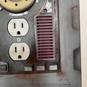 May include: A close-up of a gray, industrial-style wall plate with two white electrical outlets. The plate features metallic accents, including a circular gear-like element and a section with red bars. The overall design has a steampunk aesthetic.